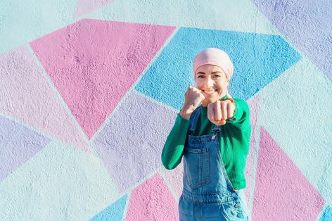 eine junge Frau mit Kopftuch steht in Kampfpose vor einer bunten Wand und lächelt