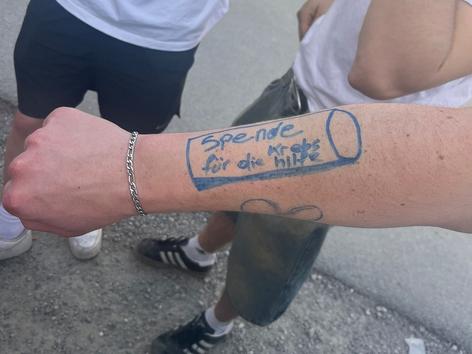 ein aufgemalter Becher auf einem Unterarm mit den Worten: Spende für die Krebshilfe