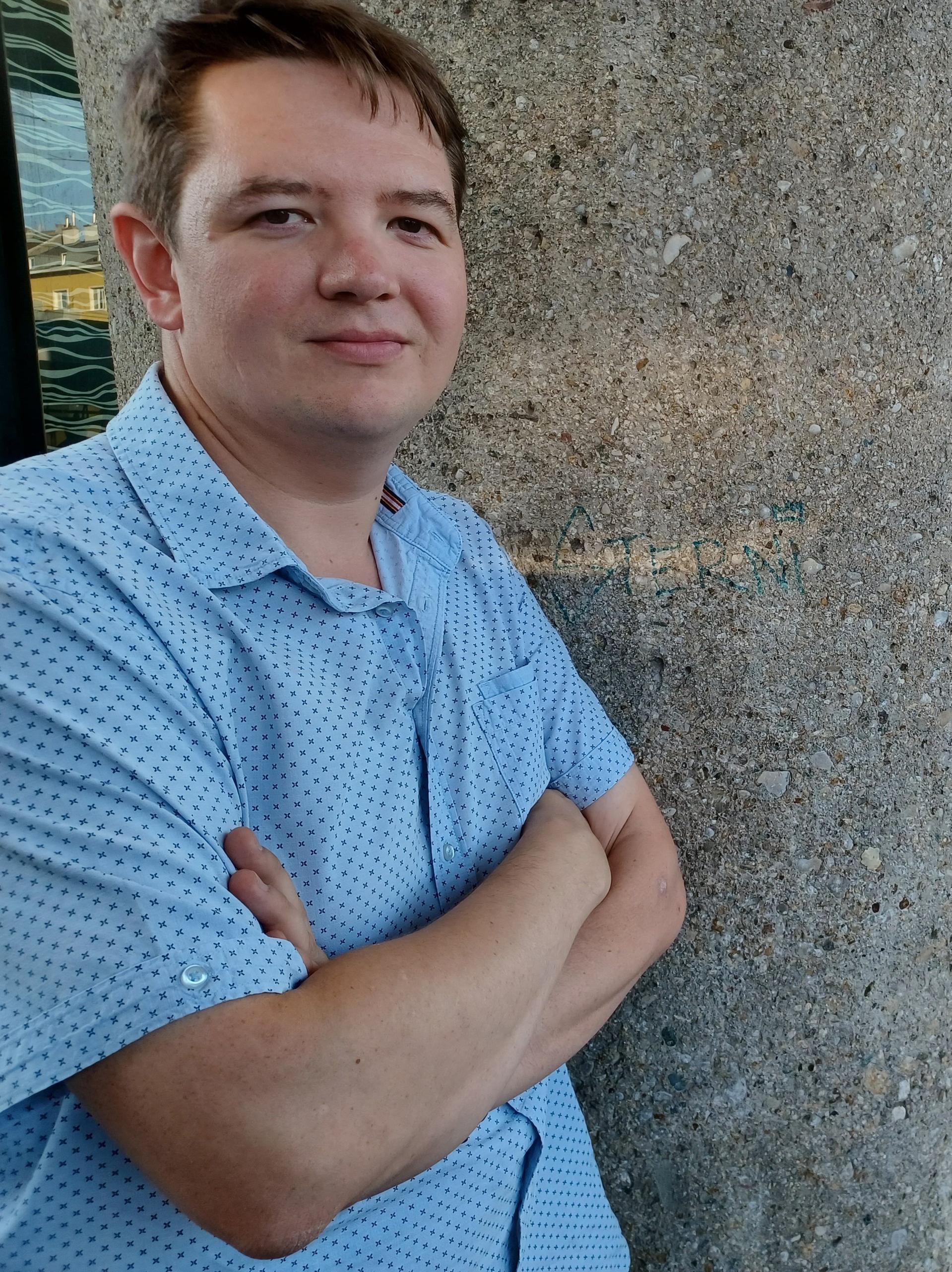 Lukas steht mit verschränkten Armen und Blick in die Kamera an eine Betonsäule gelehnt