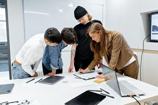 4 junge Leute stehen an Schreibtisch und stecken die Köpfe über Notizen zusammen