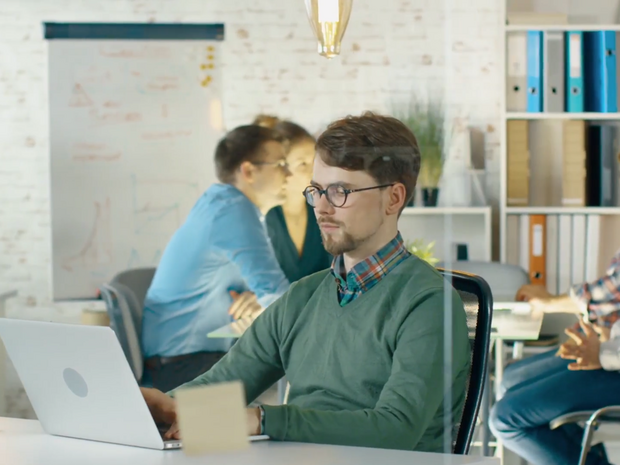 Mann arbeitet am Laptop, im Hintergrund 4 Personen bei einer Besprechung