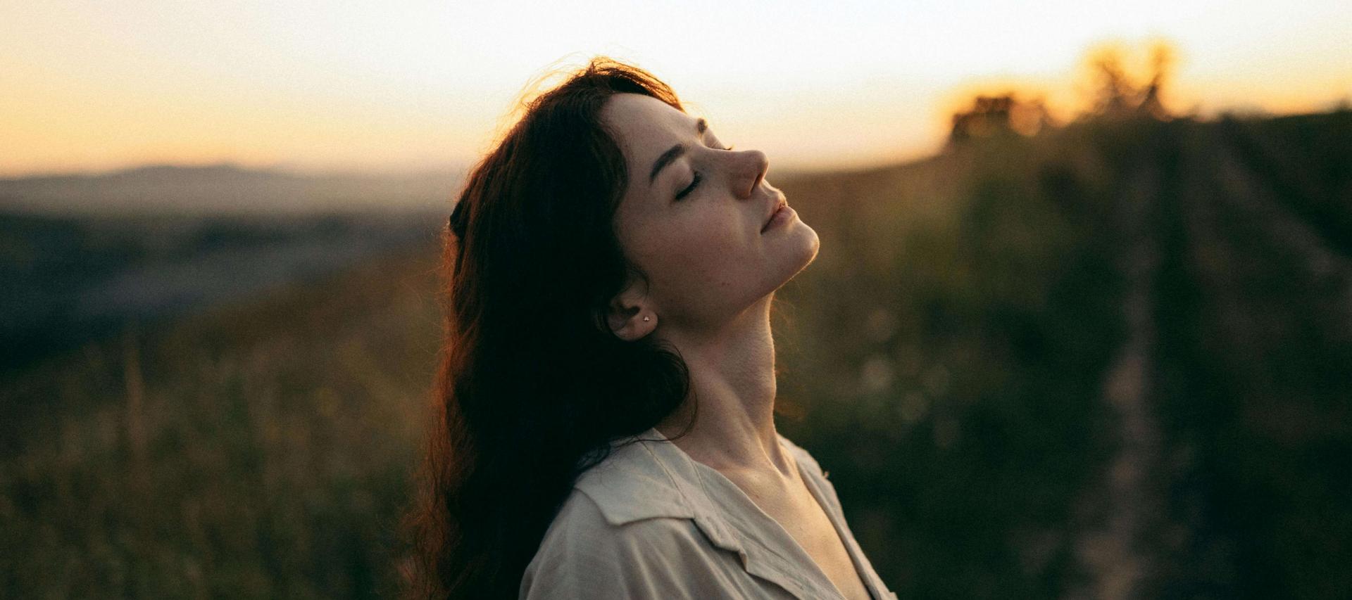 Frau steht in der Abenddämmerung den Blick Richtung Himmel gerichtet, ihre Augen sind geschlossen