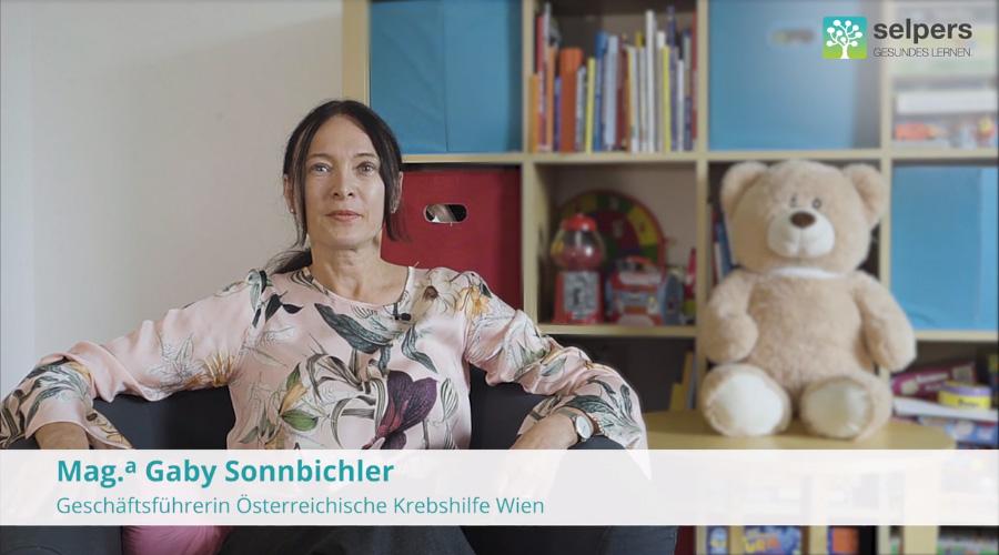 Gaby Schubert-Sonnbichler sitzt vor einem Regal mit Büchern und Kinderspielzeug, ein Teddybär im Hintergrund