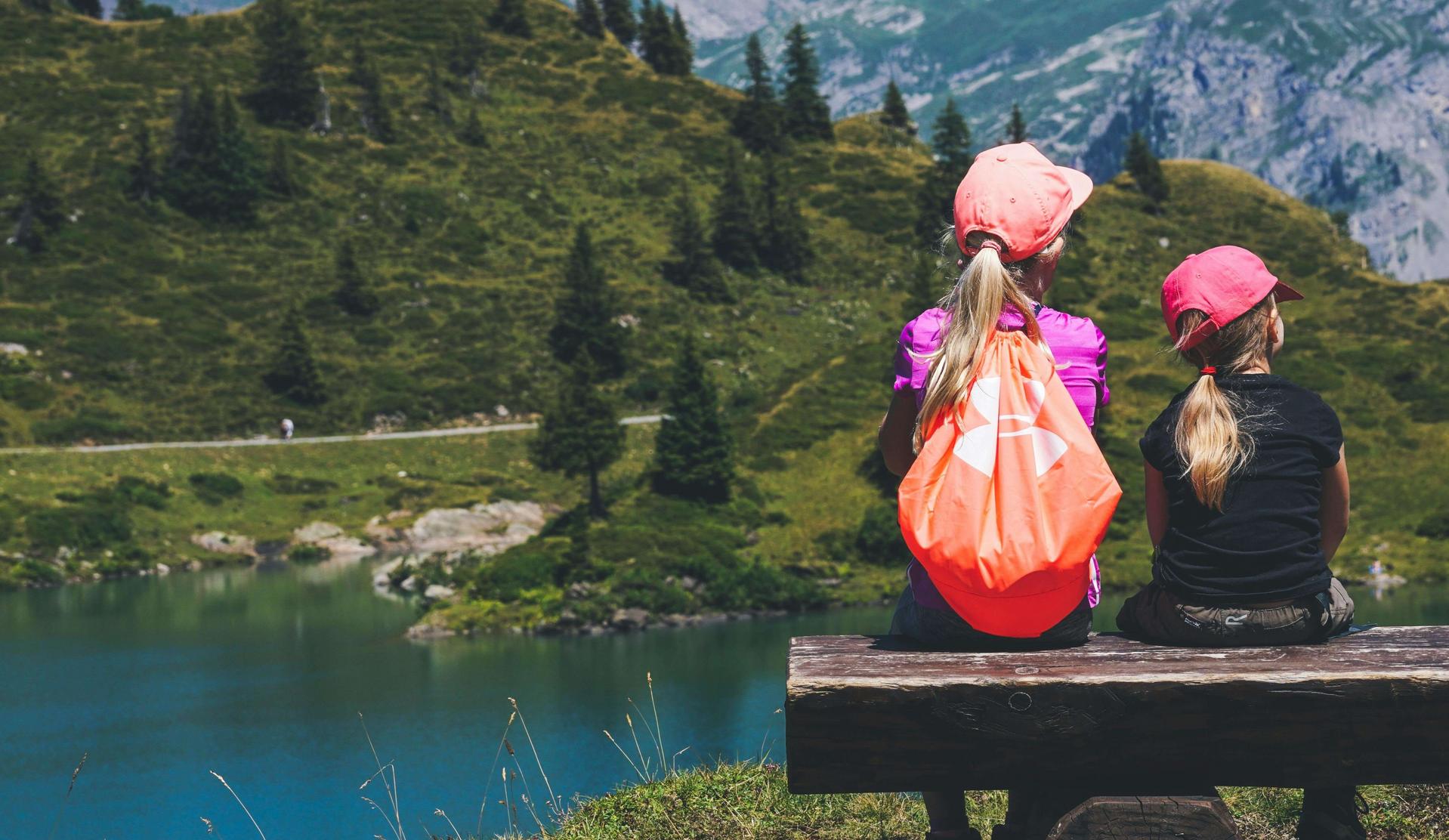 R&uuml;ckenansicht zweier M&auml;dchen, die an einem Bergsee auf einer Bank sitzen